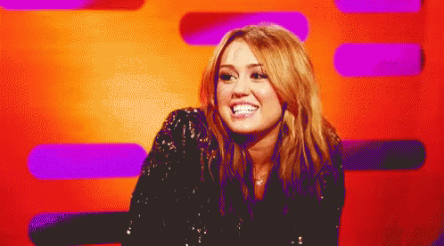 A Woman In A Black Sequined Top Smiles In Front Of A Purple And Orange Background GIF