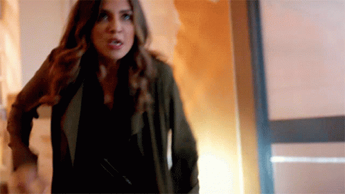 A Woman In A Black Shirt And A Green Jacket Is Standing In A Hallway . GIF