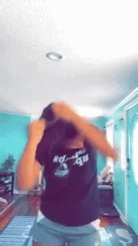 A Woman In A Black Shirt And Shorts Is Standing In A Living Room With Her Hair In A Bun . GIF