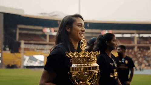 A Woman In A Black Shirt Holds A Gold Trophy With A Crown On It GIF
