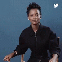 A Woman In A Black Shirt Is Sitting In A Chair With Her Hands Folded . GIF
