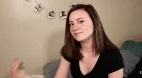 A Woman In A Black Shirt Is Sitting On A Bed In Front Of A Wall With Pictures Hanging On It . GIF