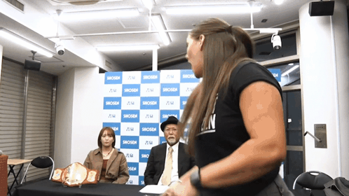 A Woman In A Black Shirt That Says ' N ' On It Stands In Front Of A Blue And White Backdrop GIF