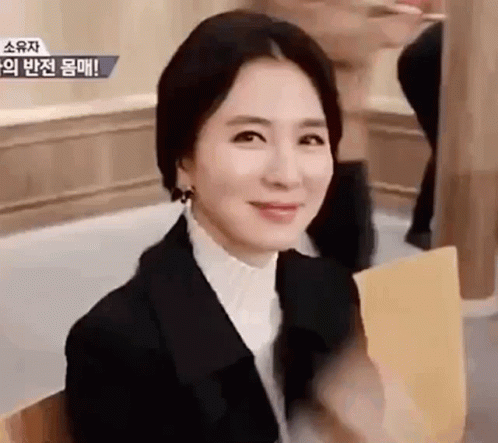 A Woman In A Black Suit And White Shirt Is Sitting In A Chair And Smiling . GIF