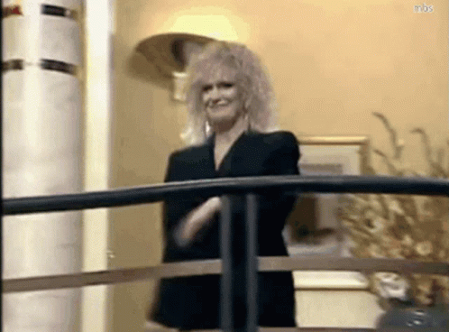A Woman In A Black Suit Is Standing On A Balcony With Her Arms Crossed . GIF