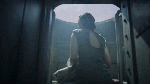 A Woman In A Black Tank Top Is Looking Out A Window GIF