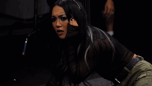 A Woman In A Black Top And Green Pants Is Kneeling Down In A Dark Room GIF