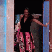A Woman In A Black Top And Pink Skirt Is Standing In A Doorway With Her Arms Outstretched . GIF