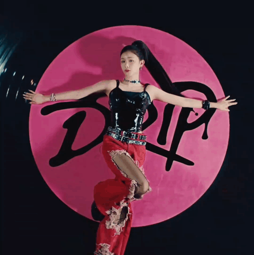 A Woman In A Black Top And Red Pants Stands In Front Of A Pink Circle That Says Drip GIF