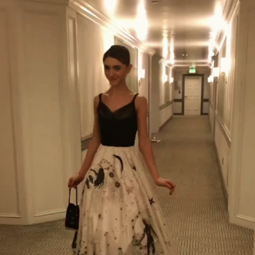 A Woman In A Black Top And White Skirt Is Walking Down A Hallway GIF