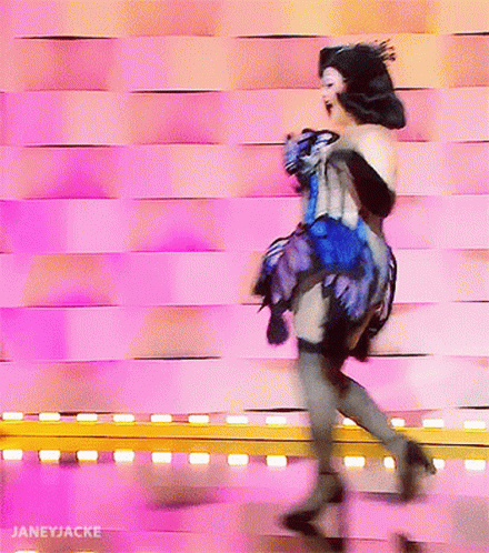A Woman In A Blue And Black Dress Is Dancing In Front Of A Pink Wall With Janeyjacke Written On The Bottom GIF