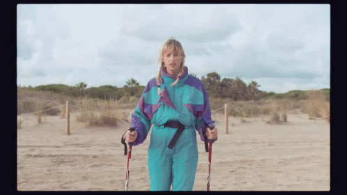 A Woman In A Blue And Purple Ski Suit Holds Walking Poles GIF