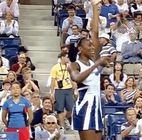 A Woman In A Blue And White Dress Is Raising Her Fist In The Air In Front Of A Crowd GIF