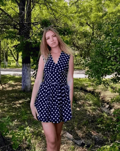 A Woman In A Blue And White Polka Dot Dress Stands In A Park GIF