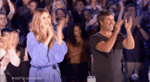 A Woman In A Blue Dress And A Man In A Black Shirt Are Applauding In Front Of A Crowd . GIF