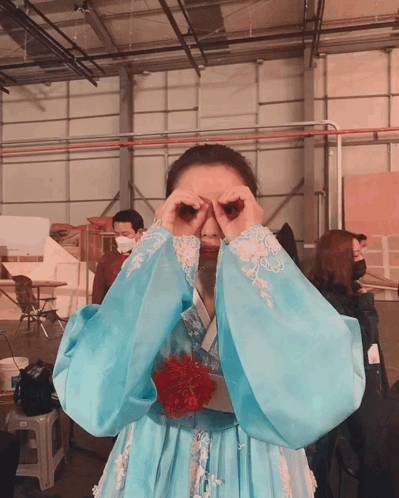 A Woman In A Blue Dress Covering Her Eyes With Her Hands In A Warehouse GIF