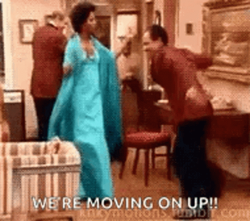 A Woman In A Blue Dress Is Dancing In A Living Room With A Man In A Suit . GIF