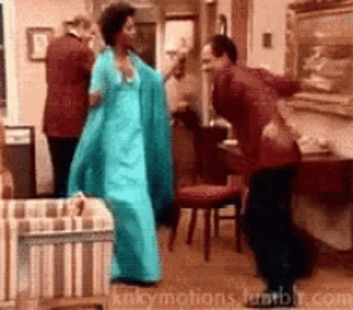 A Woman In A Blue Dress Is Dancing With A Man In A Red Suit In A Living Room GIF