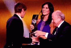 A Woman In A Blue Dress Is Holding A Trophy And Shaking Hands With A Man In A Suit GIF