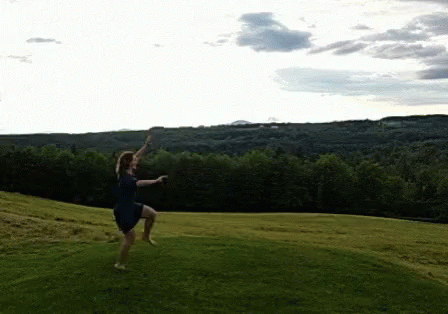 A Woman In A Blue Dress Is Jumping In The Air In A Grassy Field GIF