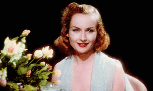 A Woman In A Blue Dress Is Smiling In Front Of A Vase Of Flowers GIF