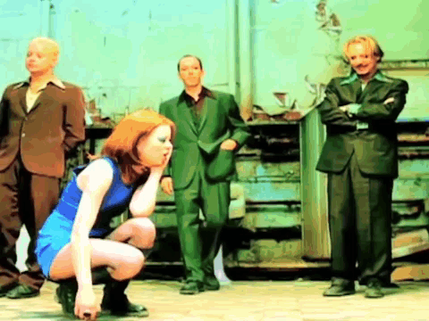 A Woman In A Blue Dress Is Squatting Down In Front Of Three Men In Suits GIF