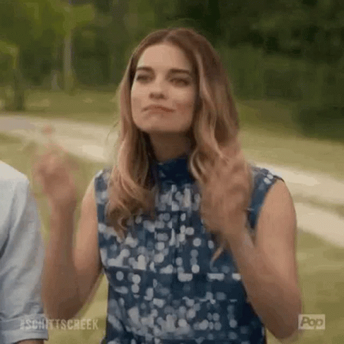 A Woman In A Blue Dress Is Standing In A Park With Her Hands In The Air . GIF