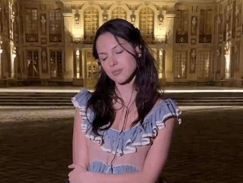 A Woman In A Blue Dress Is Standing In Front Of A Building With Her Eyes Closed GIF