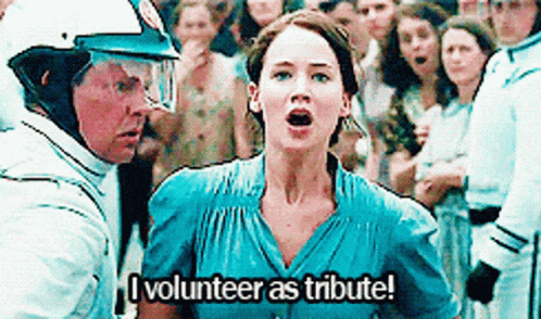 A Woman In A Blue Dress Is Standing In Front Of A Crowd Of People And Says `` I Volunteer As Tribute ! '' GIF