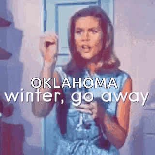 A Woman In A Blue Dress Is Standing In Front Of A Door Holding A Microphone And Saying Oklahoma Winter Go Away . GIF