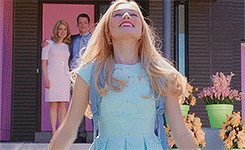 A Woman In A Blue Dress Is Standing In Front Of A Pink House With Her Arms Outstretched . GIF