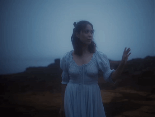 A Woman In A Blue Dress Stands On A Rocky Shore GIF