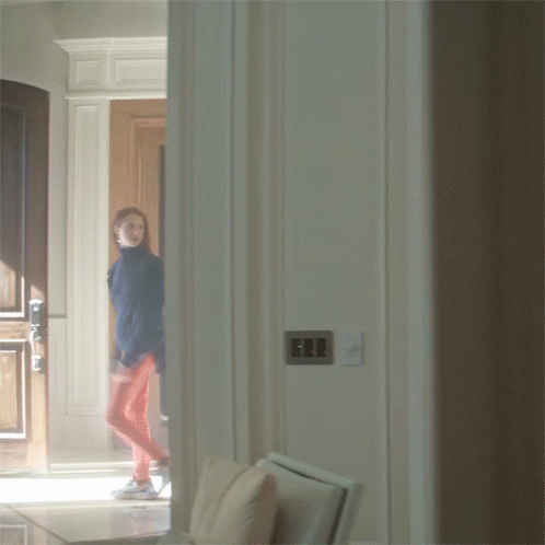 A Woman In A Blue Jacket And Red Pants Is Walking Through A Doorway GIF