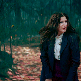 A Woman In A Blue Jacket And White Shirt Is Walking Through A Forest GIF
