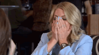 A Woman In A Blue Jacket Is Covering Her Face With Her Hands While Sitting At A Table . GIF