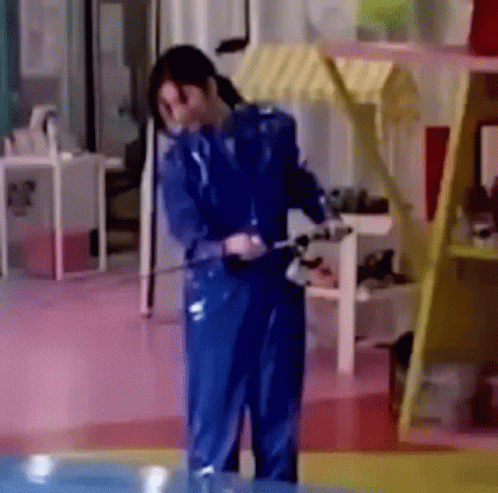 A Woman In A Blue Jumpsuit Is Standing In A Room With A Hose . GIF