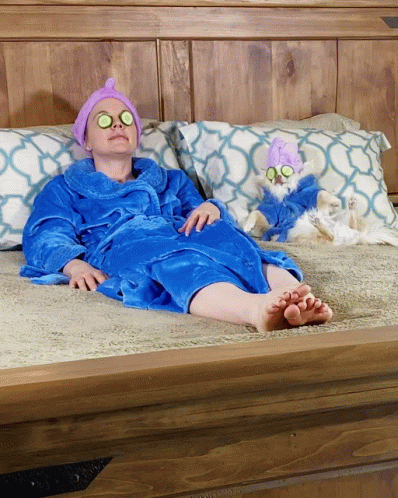 A Woman In A Blue Robe Is Laying On A Bed Next To A Small White Dog GIF