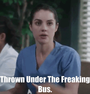 A Woman In A Blue Scrub Is Sitting In Front Of A Sign That Says " Thrown Under The Freaking Bus " GIF