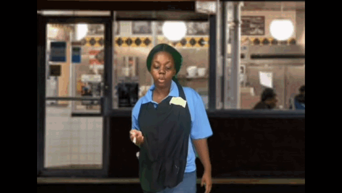 A Woman In A Blue Shirt And Black Apron Is Standing In Front Of A Store GIF
