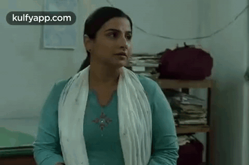 A Woman In A Blue Shirt And White Scarf Is Standing In A Room With A Stack Of Books On A Shelf . GIF