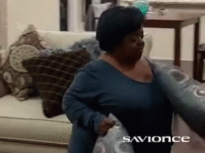 A Woman In A Blue Shirt Is Holding A Pillow In Front Of A Sign That Says " Savonce " GIF
