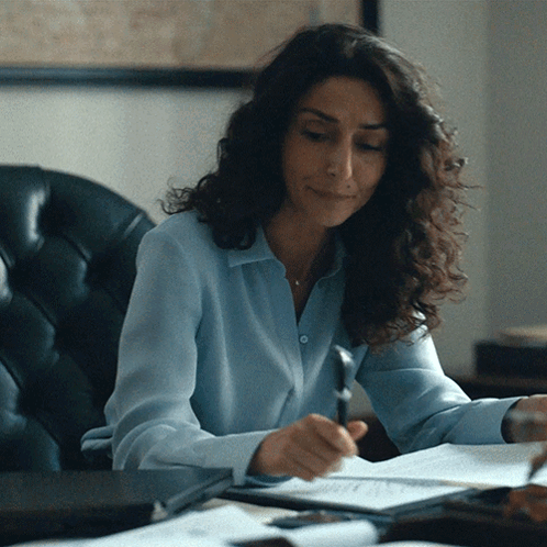A Woman In A Blue Shirt Is Sitting At A Desk Writing On A Piece Of Paper GIF