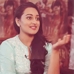 A Woman In A Blue Shirt Is Sitting In Front Of A Painting And Talking To Someone . GIF