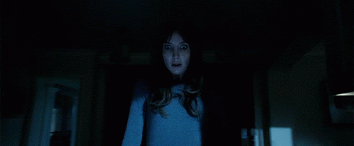 A Woman In A Blue Shirt Is Standing In A Dark Room Looking At The Camera GIF