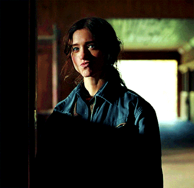 A Woman In A Blue Shirt Is Standing In A Dark Room Looking At The Camera GIF