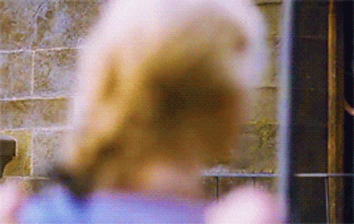 A Woman In A Blue Shirt Is Standing In Front Of A Brick Wall Looking Out A Window . GIF
