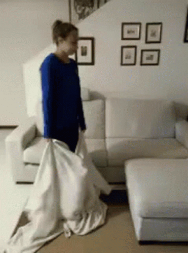 a woman in a blue shirt is standing in front of a white couch holding a white sheet