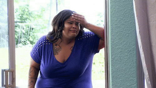 A Woman In A Blue Shirt Is Standing In Front Of A Window And Covering Her Forehead With Her Hand . GIF