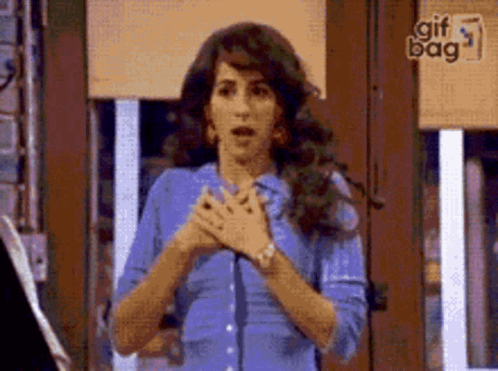 A Woman In A Blue Shirt Is Standing In Front Of A Window With Her Hands Folded In Front Of Her Chest . GIF