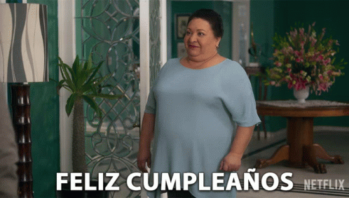 A Woman In A Blue Shirt Stands In Front Of A Sign That Says " Feliz Cumpleanos " GIF